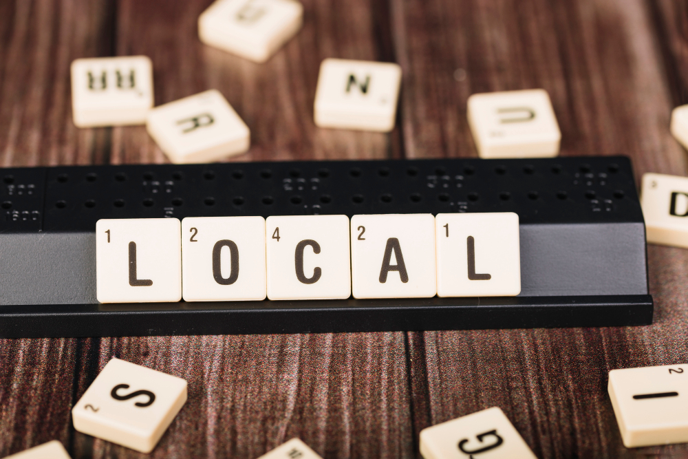 Scrabble tiles spell out "LOCAL" on a wooden surface with scattered letters.